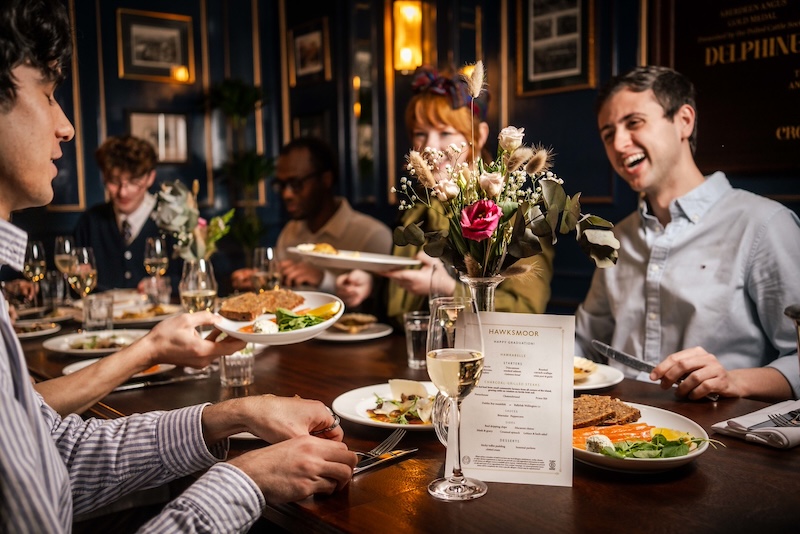A shot of a private dining event, with smartly dressed guests in shirts around the dining table, smiling and laughing with plates of food and champaign on the table in front of them.