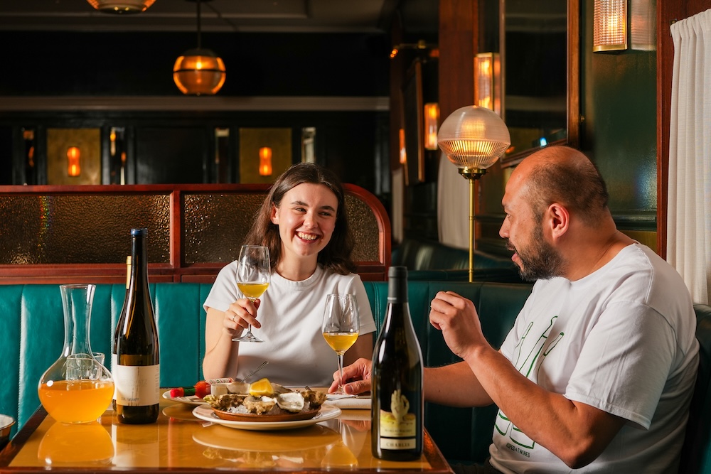 A couple sat at a dining table in a steakhouse, holding glasses of wine and laughing. On the table are plates of seafood and bottles of white wine.