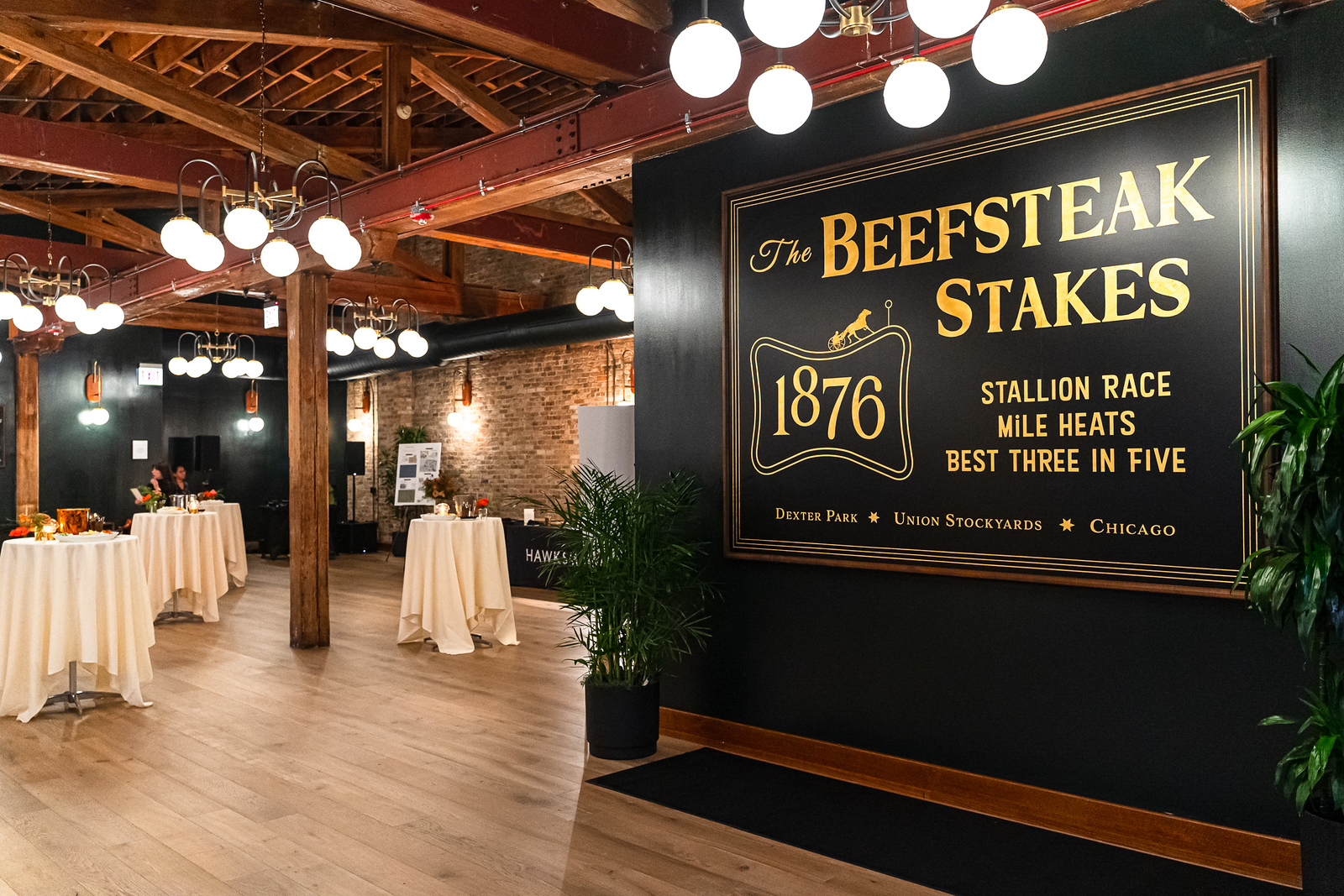 A wide shot of a large, high ceiling private function room, with 3 poseur tables covered in white table cloths, with floral arrangements