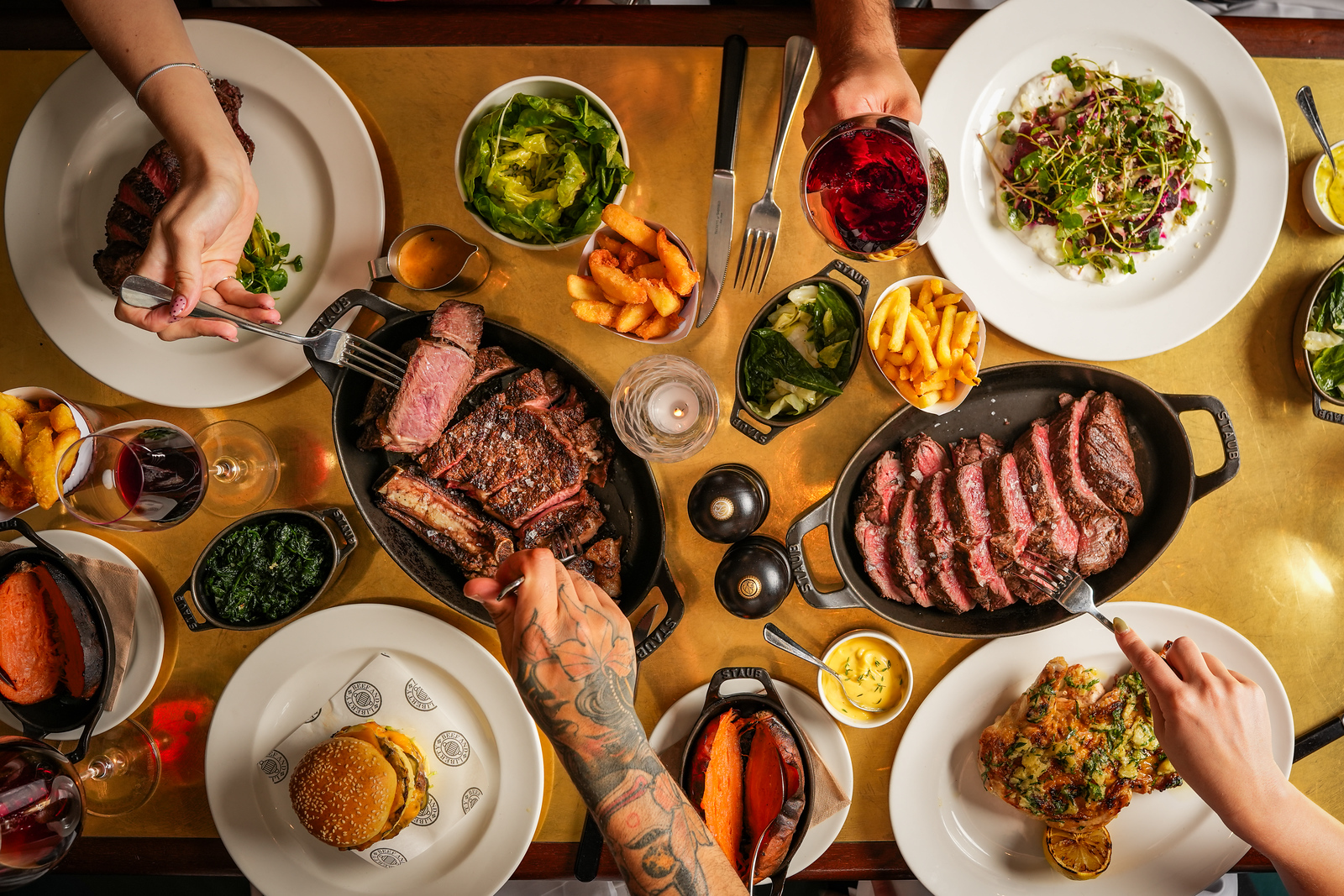 An overhead shot of a golden dining table laden with steaks, sides and starters, with hands reaching out to pick up the dishes