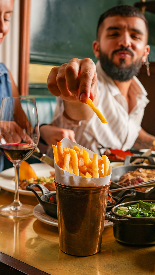 A young smiling man reaching out over the table to grab a fry from a copper pot