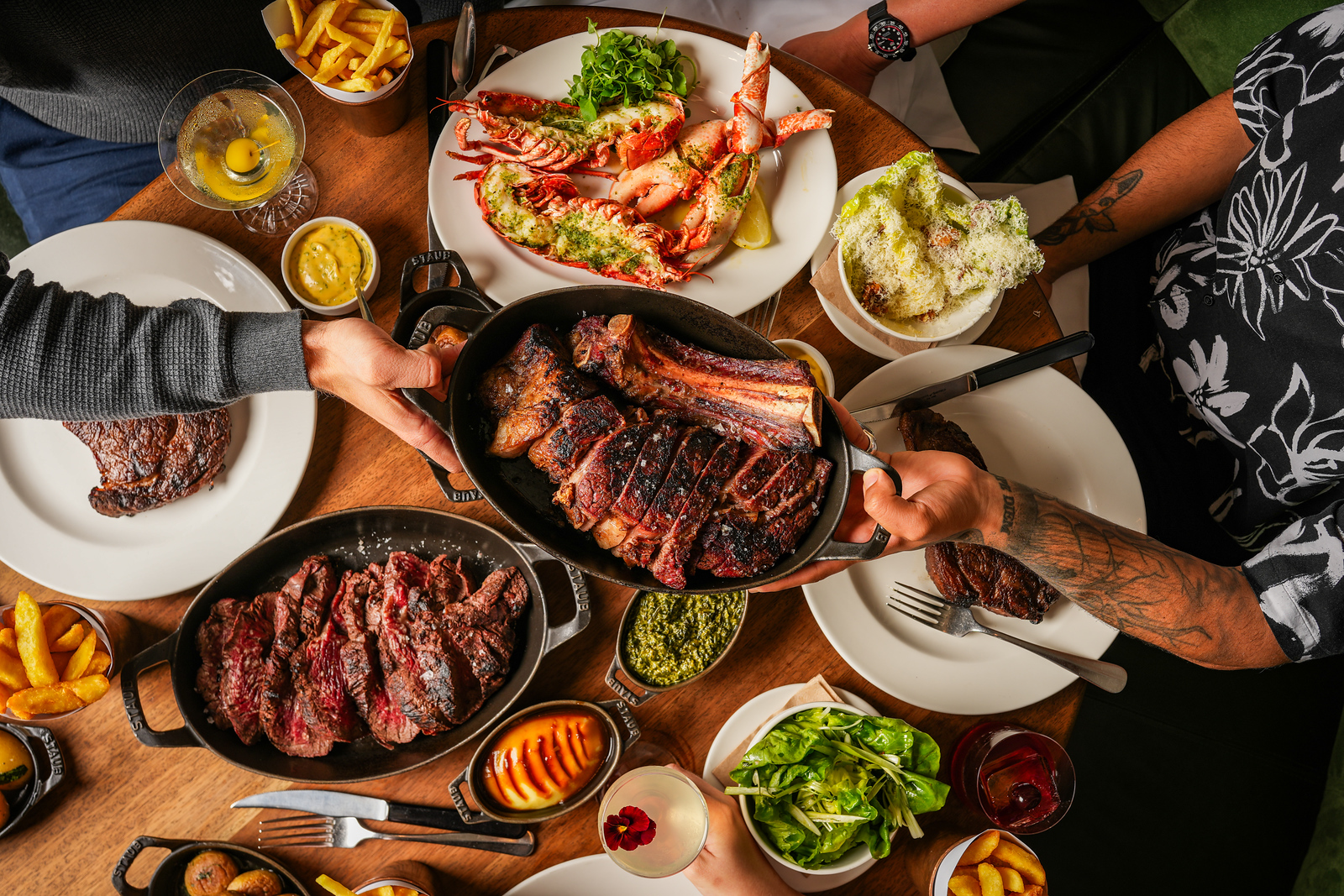 An overhead shot of a table generously laden with steaks and seafood; a skillet with a sharing steak being passed in the foreground.