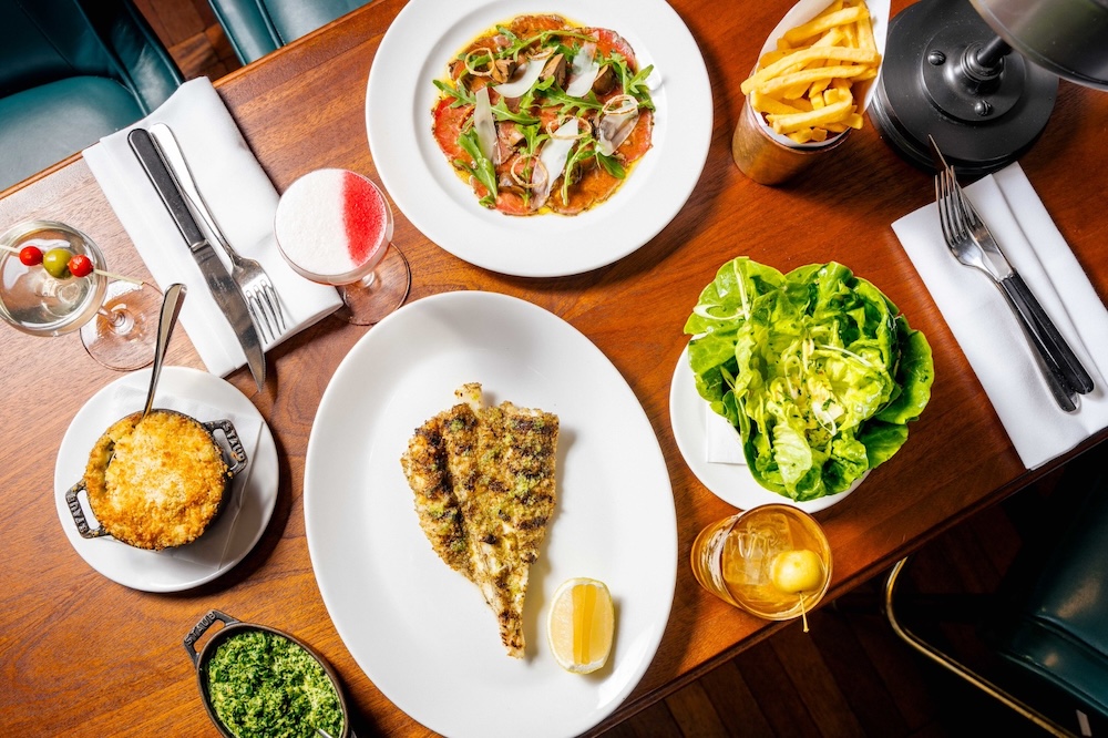 An overhead shot of a wooden table with plates of crudo, monkfish and a side dish of mac and cheese.