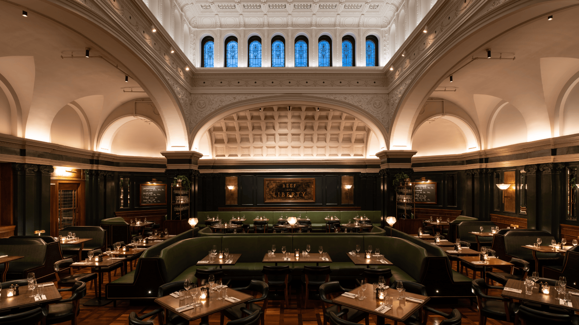 A wide angle shot of Hawksmoor New York dining room - stately room under the high vaulted ceilings, with green leather banquette seating and tables