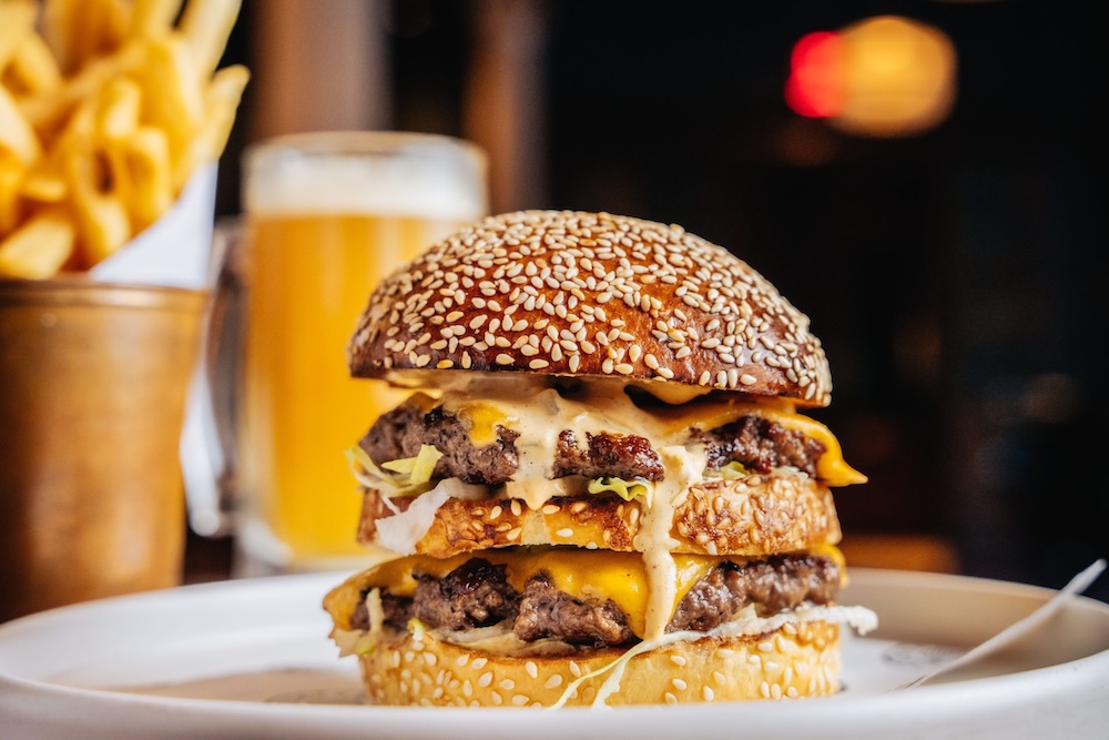 The Notorious BEEF burger, which includes two smash burger patty's and cheese dripping down onto the plate. In the background a serving of fries and a cocktail are visible but slightly out of focus.