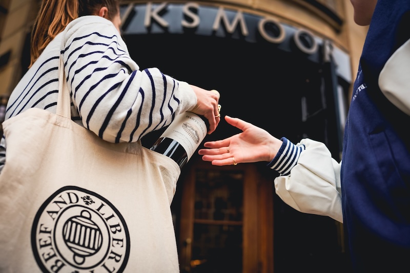 Two women outside a Hawksmoor restaurant. One of them is carrying a bottle of wine in a canvas bag. She is about to pass it to the outstretched hand of the other woman.