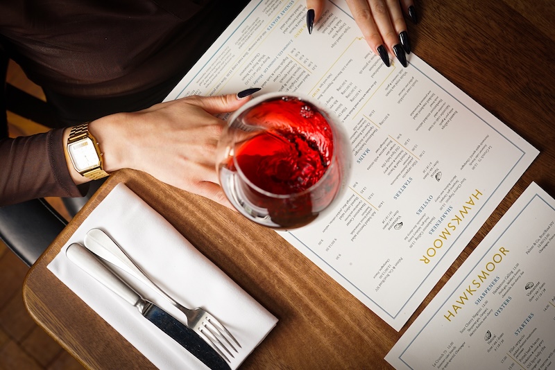 a glass of red placed on top of a food menu with Hawksmoor logo, on a wooden table. A woman's hands are resting on the menu. To the right of her is a knife and folk and a napkin.