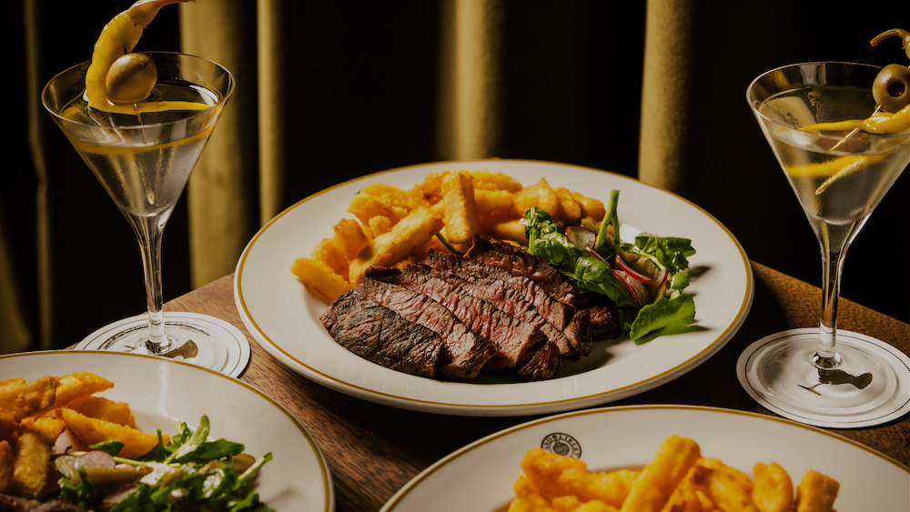 Rump steak sliced up, with a side of fries, on a white plate against an olive green curtain. The steak is surrounded by more plates of steak and two martinis garnished with an olive either side of the plate of steak.