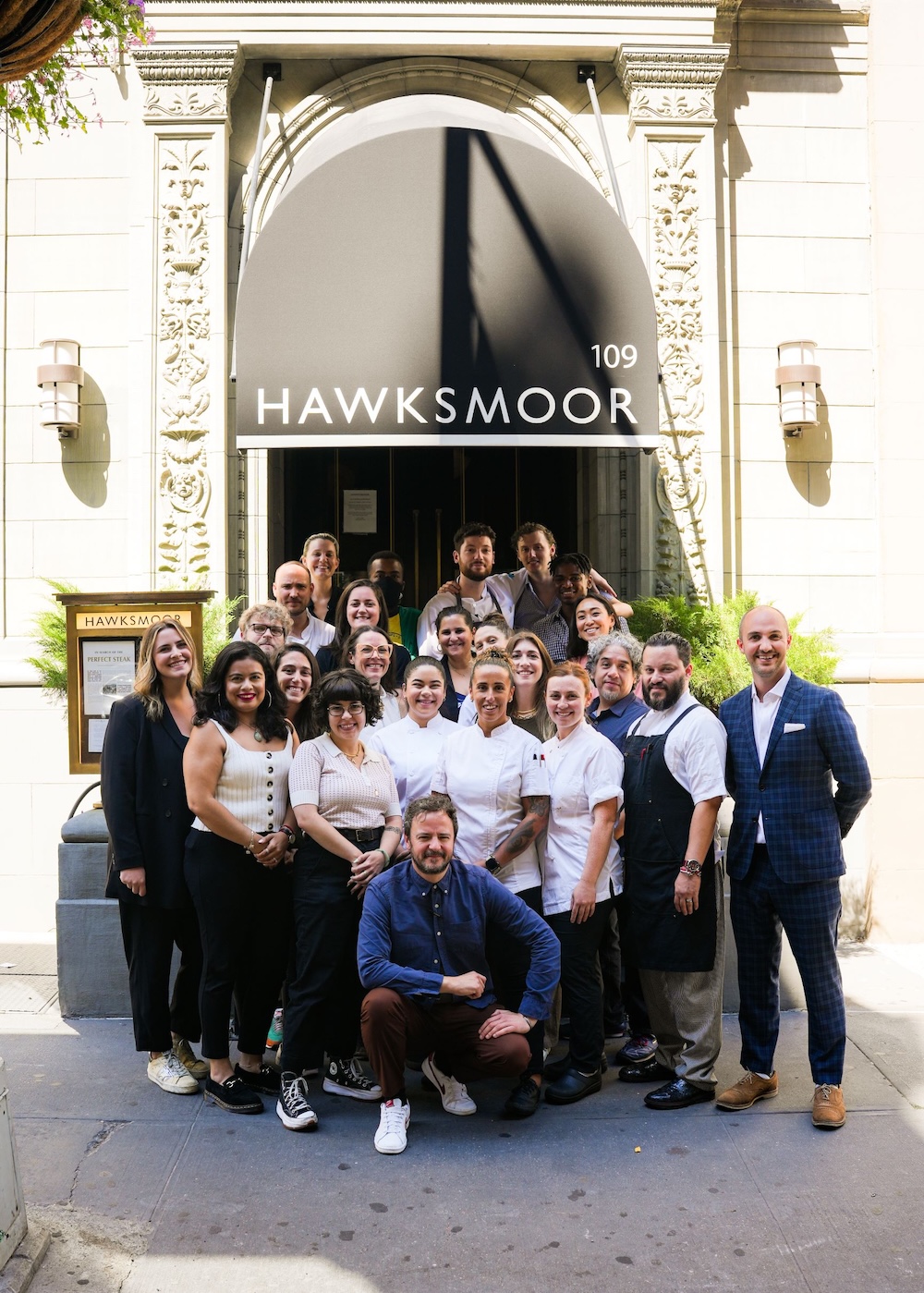 A shot of full team outside Hawksmoor New York on a sunny day. They are collected together as a group in their work attire, some in chef's whites and some in neat shirts, smiling at the camera.