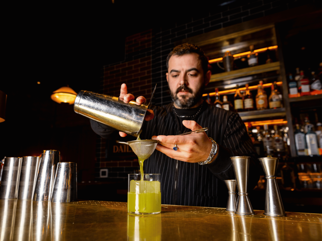 A bartender pours a bright green cocktail through a strainer into a glass at a dimly lit bar, with liquor bottles and metal shakers on the counter.