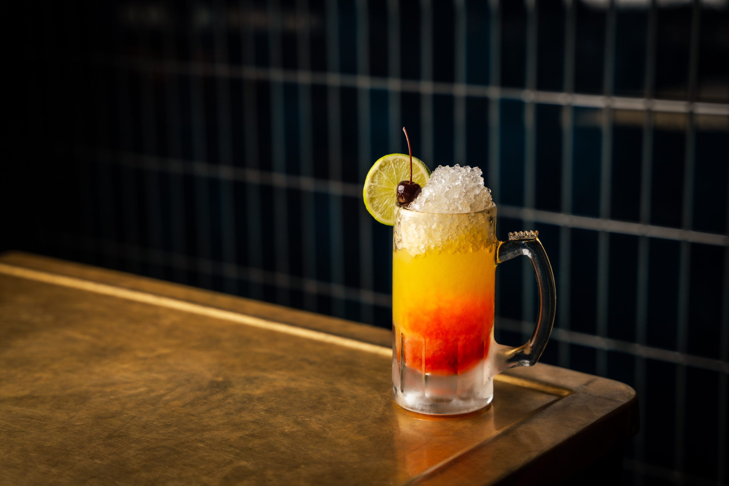 A layered tropical cocktail in a handled glass mug, topped with crushed ice, a lime slice, and a cherry on a brass table.