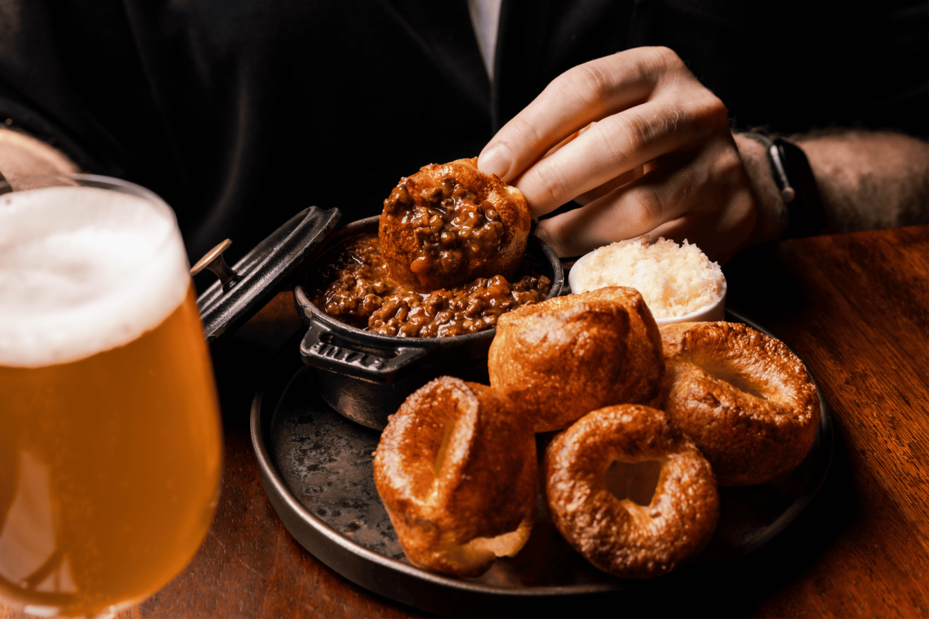 A person dips a golden Yorkshire pudding into a small cast iron pot of rich, minced meat gravy, with more Yorkshire puddings, a dish of grated horseradish, and a hazy pint of beer on a wooden table.