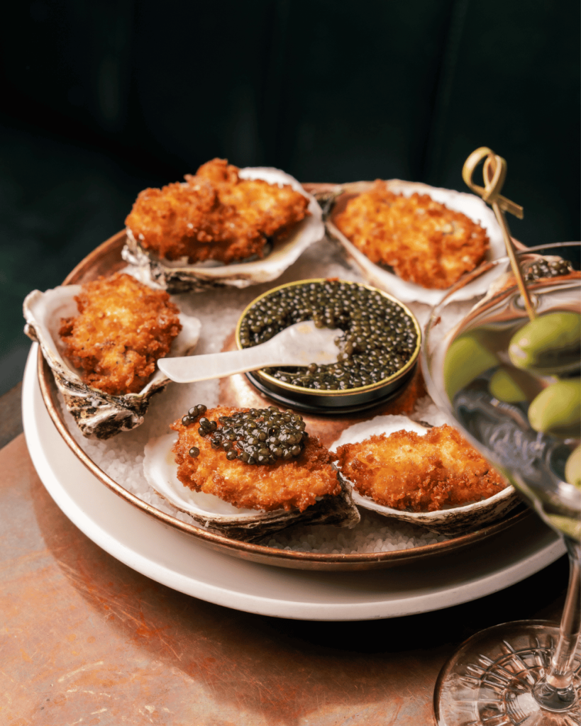 New ALC Menu 2 Five deep fried oysters in shells arranged on a bed of salt around a central tin of black caviar, with a martini glass visible in the foreground.