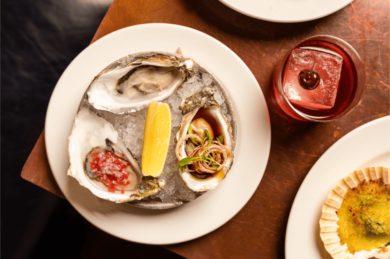 Chicago 13 A top-down view of three oysters on the half shell served on crushed ice in a round dish, garnished with a lemon wedge and different toppings, with a red cocktail on the side and part of a baked scallop dish visible on neighboring plates.