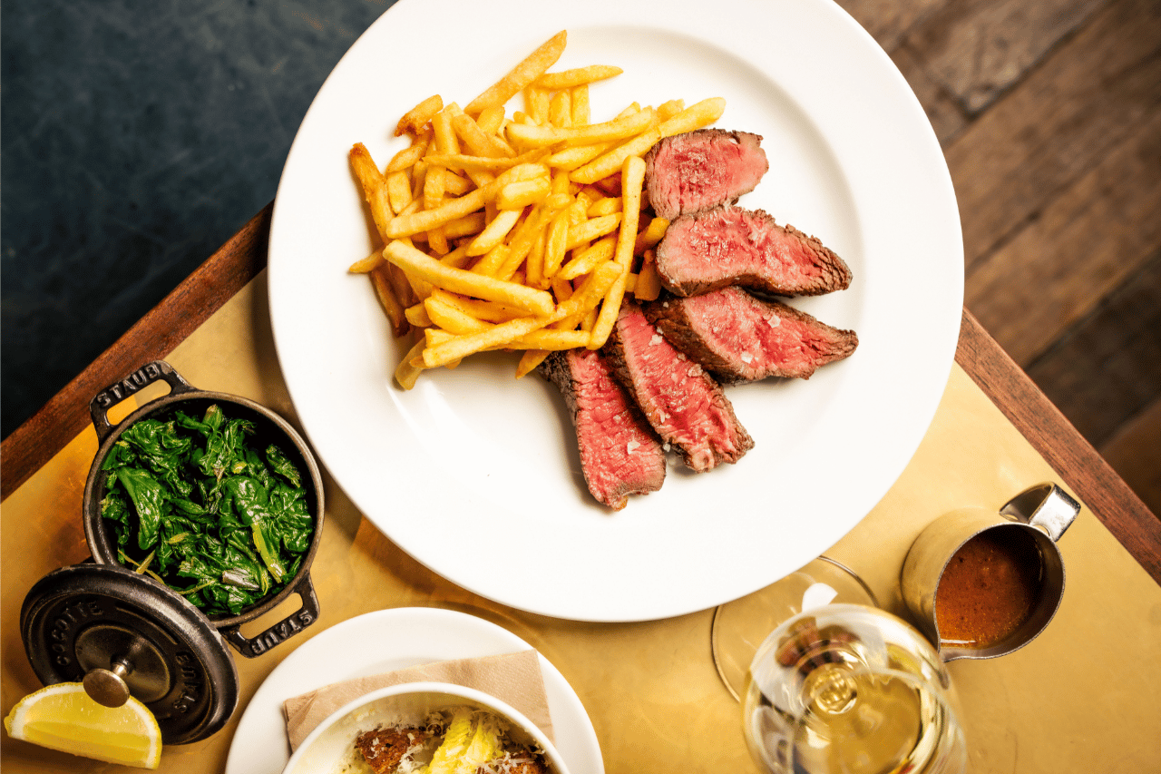 Chicago 14 A top-down view of a sliced medium-rare steak with French fries on a white plate, accompanied by a small pot of sautéed greens, a ramekin dish, a glass of white wine, and a small metal jug of sauce on a brass-topped table.