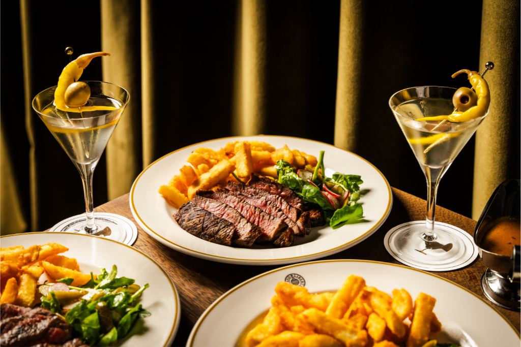 Seared steak with golden fries, mixed green salad and three martinis garnished with olives and peppers on a wooden restaurant table.