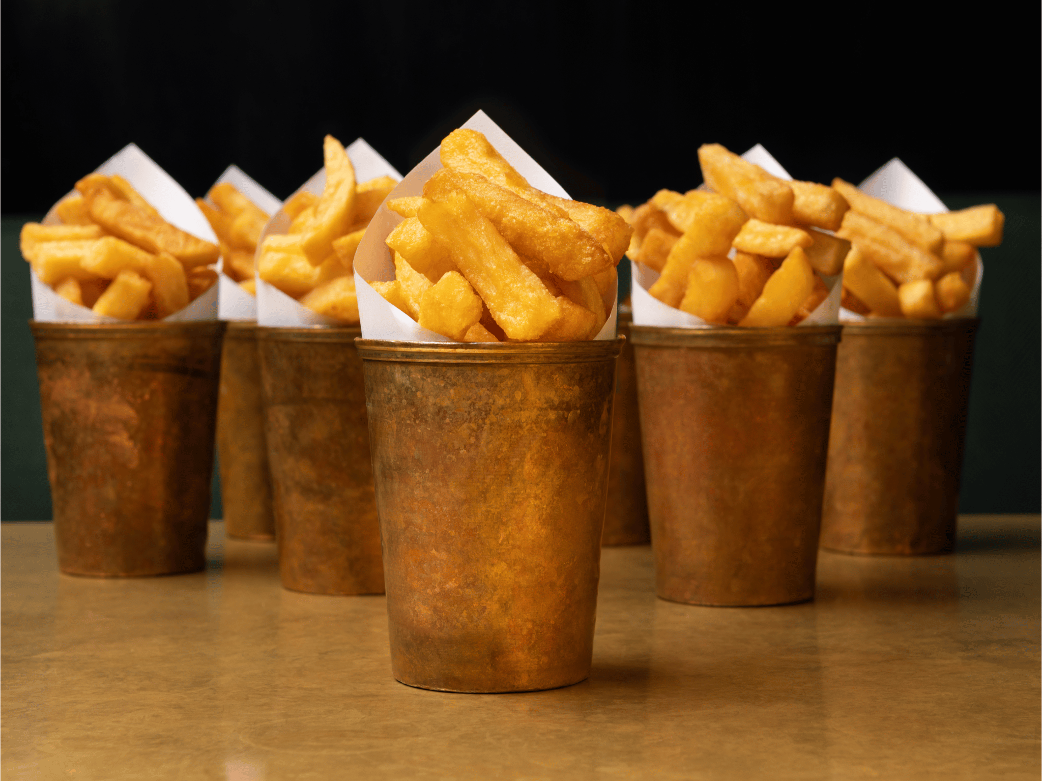 What's On 3 Five copper cups filled with thick, golden triple-cooked chips, each cup lined with white paper, arranged on a brown tabletop.