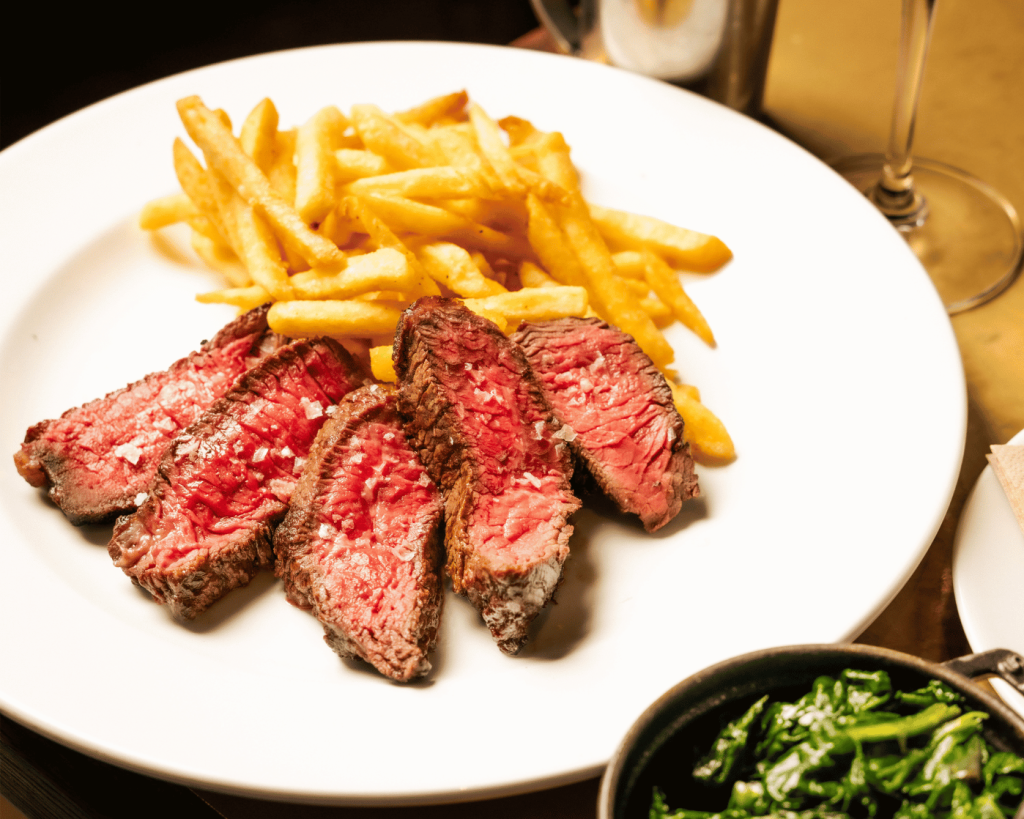 A white plate with sliced medium rare steak sprinkled with salt, served alongside a pile of thin golden fries, with a small black dish of sautéed greens partially visible in the foreground.