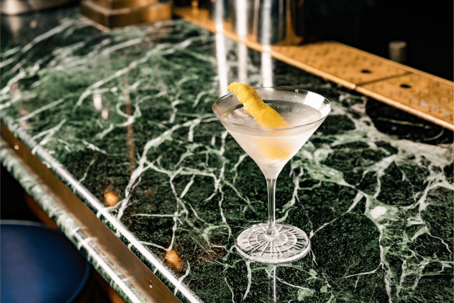A classic martini garnished with a lemon twist, served in a stemmed glass on a polished green marble bar counter.
