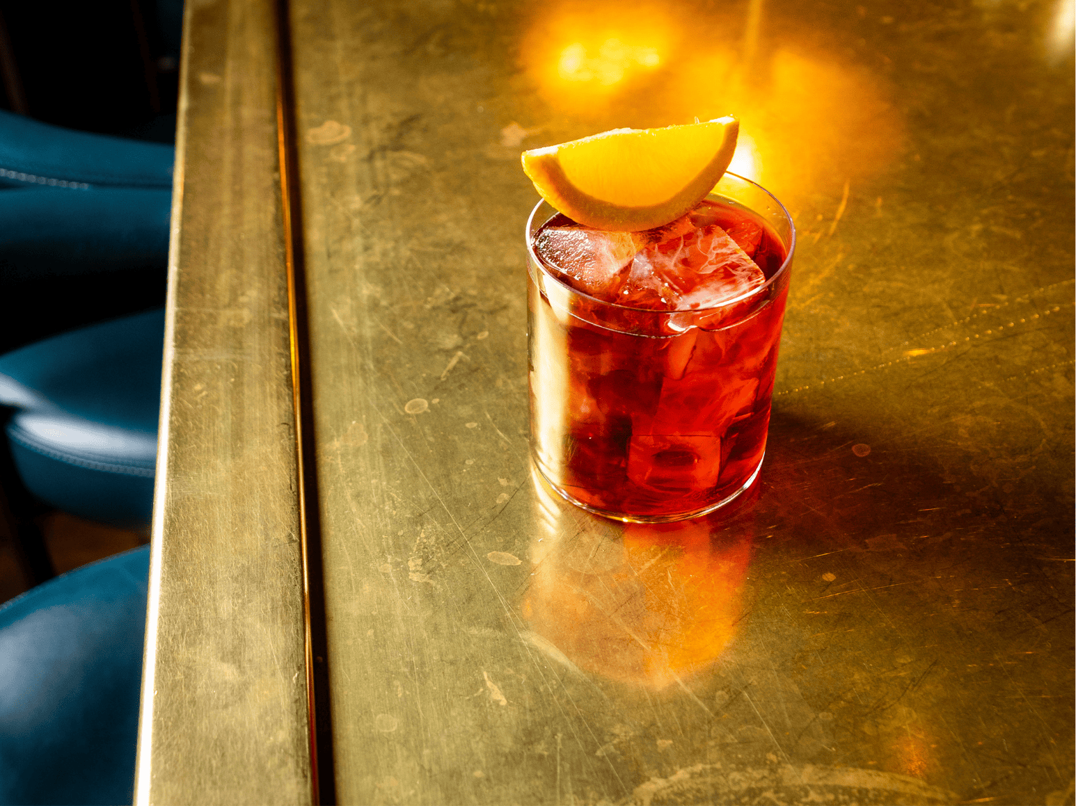 A short tumbler glass of red cocktail with ice and an orange wedge garnish, sitting on a reflective golden bar counter with soft warm lighting.