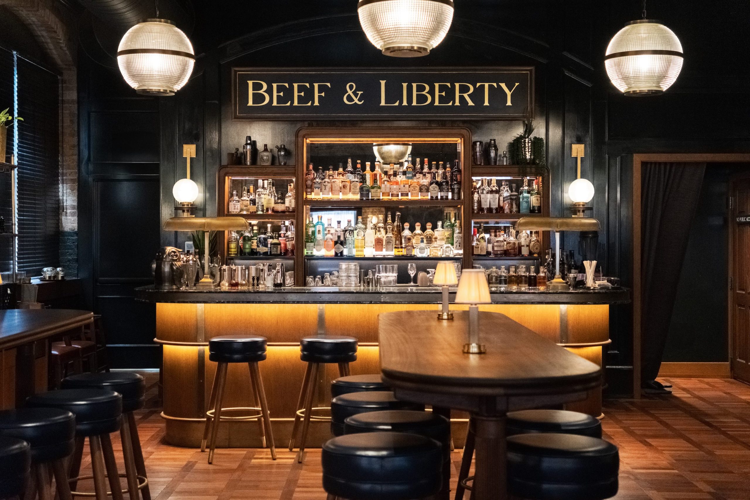 "Beef & Liberty" bar area featuring blue stools, a marble counter, wood tables, small lamp lights, and a backdrop of spirits and glassware.