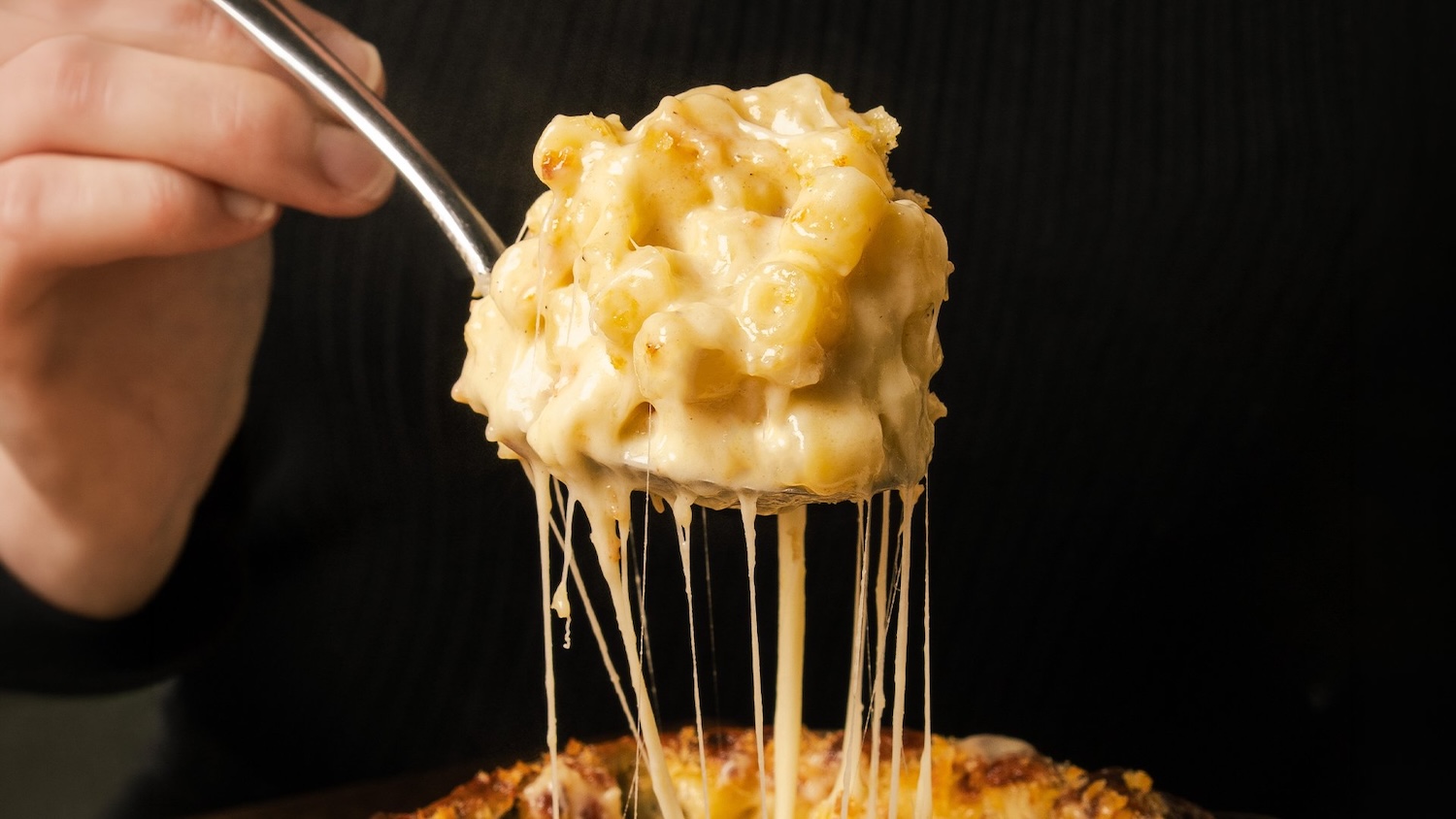 A big spoonful of mac and cheese, against a black background. There are lots of strings of cheese visible as the spoon is lifted up.