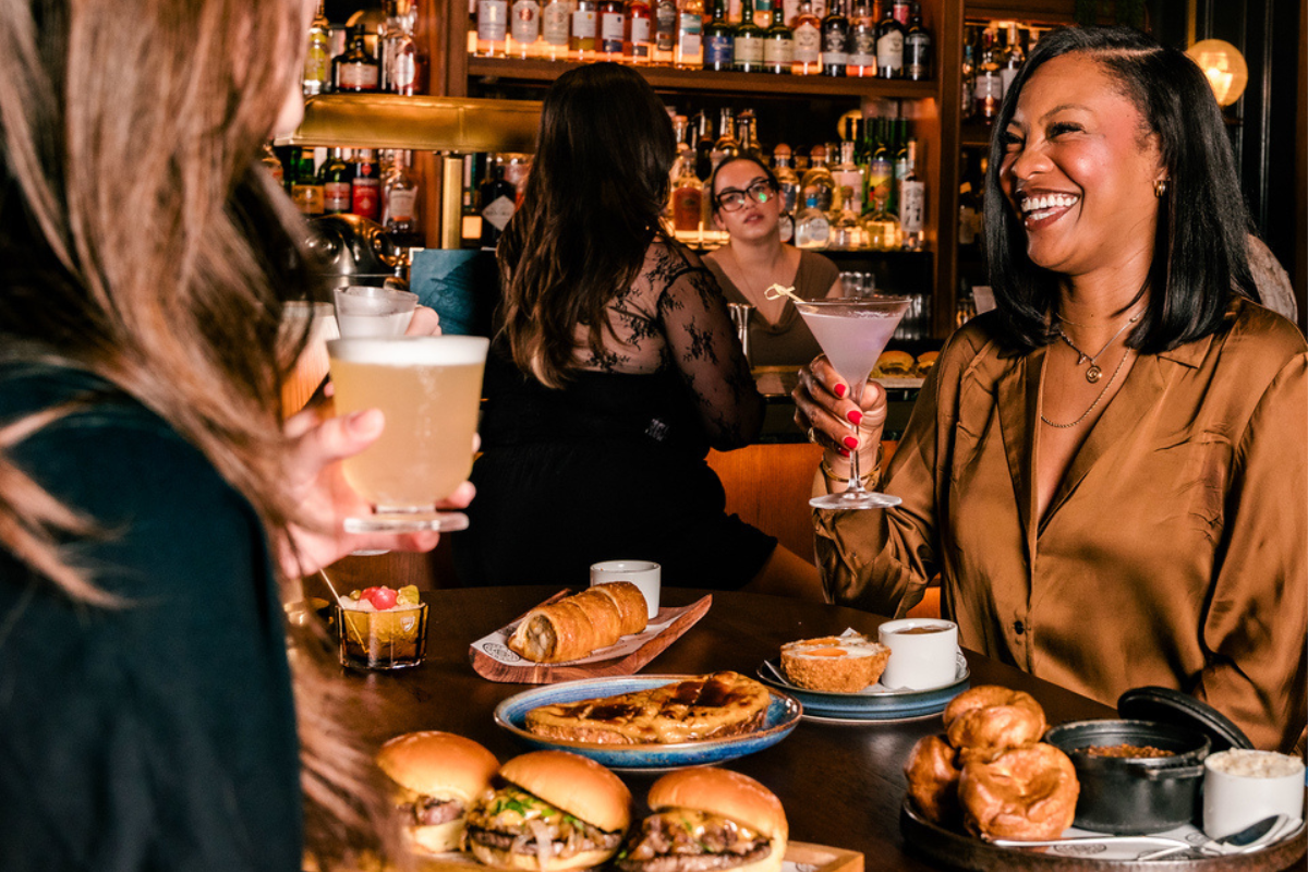 A woman smiling at her friend, holding a martini; the table is laden with sliders and bar snacks