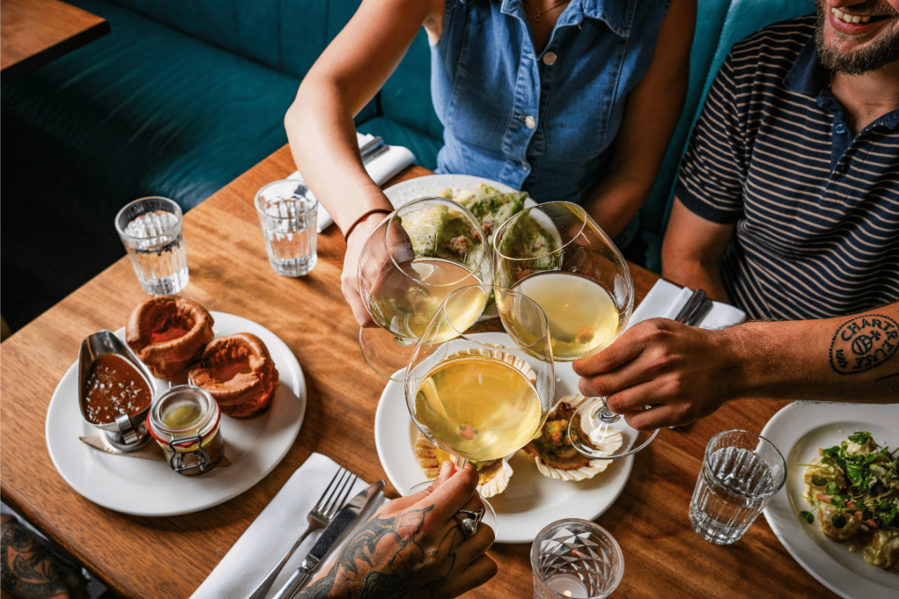 MAKE JANUARY JOYOUS - hawksmoor liverpool 2 Top-down view of three people clinking large glasses of white wine over a wooden restaurant table set with plates of food, including Yorkshire puddings with gravy, scallop shells, and a salad, alongside water glasses and neatly folded napkins with cutlery.