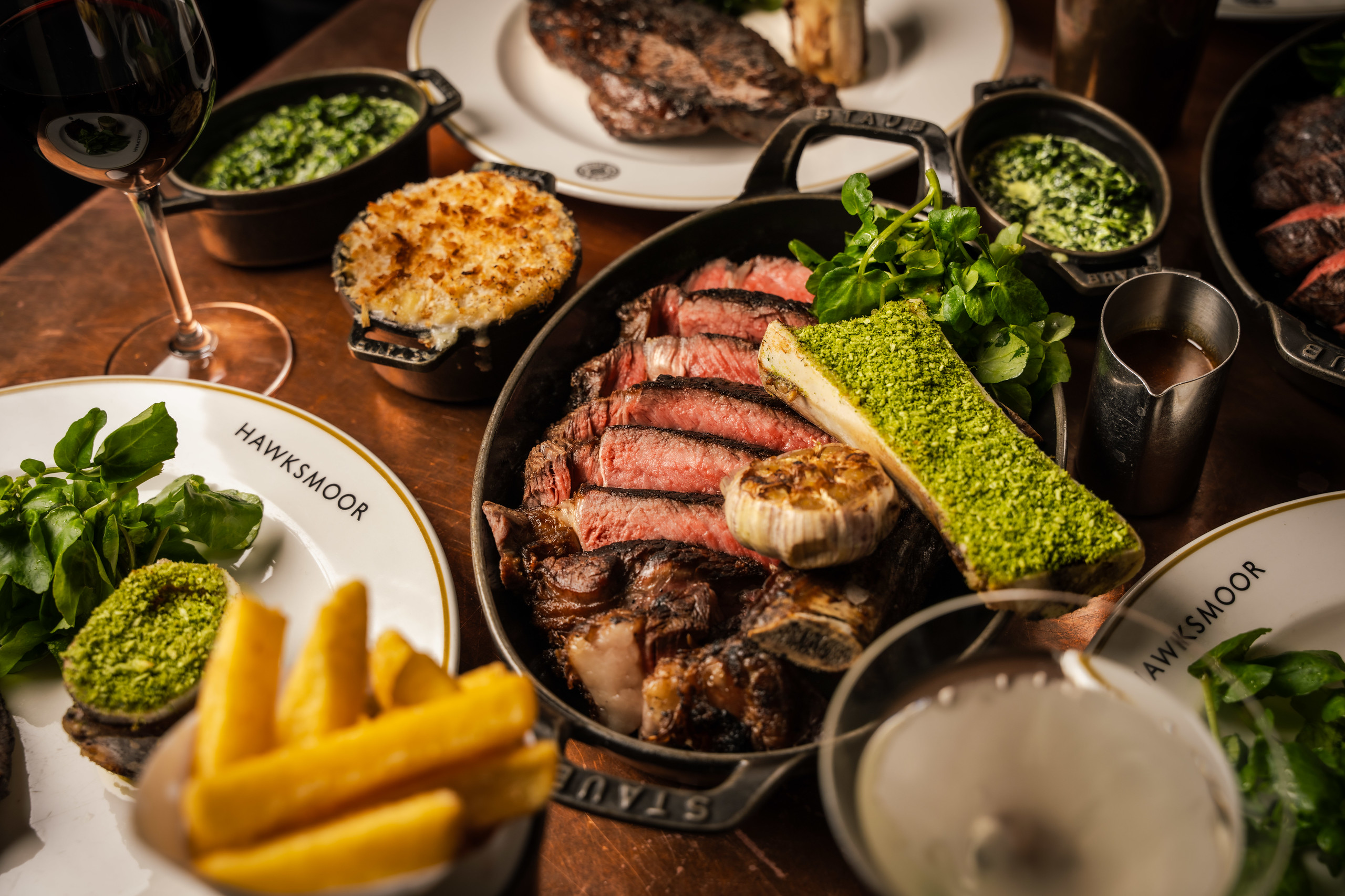 A skillet with a sharing Longhorn steak on a wooden table, surrounded by side dishes and drinks