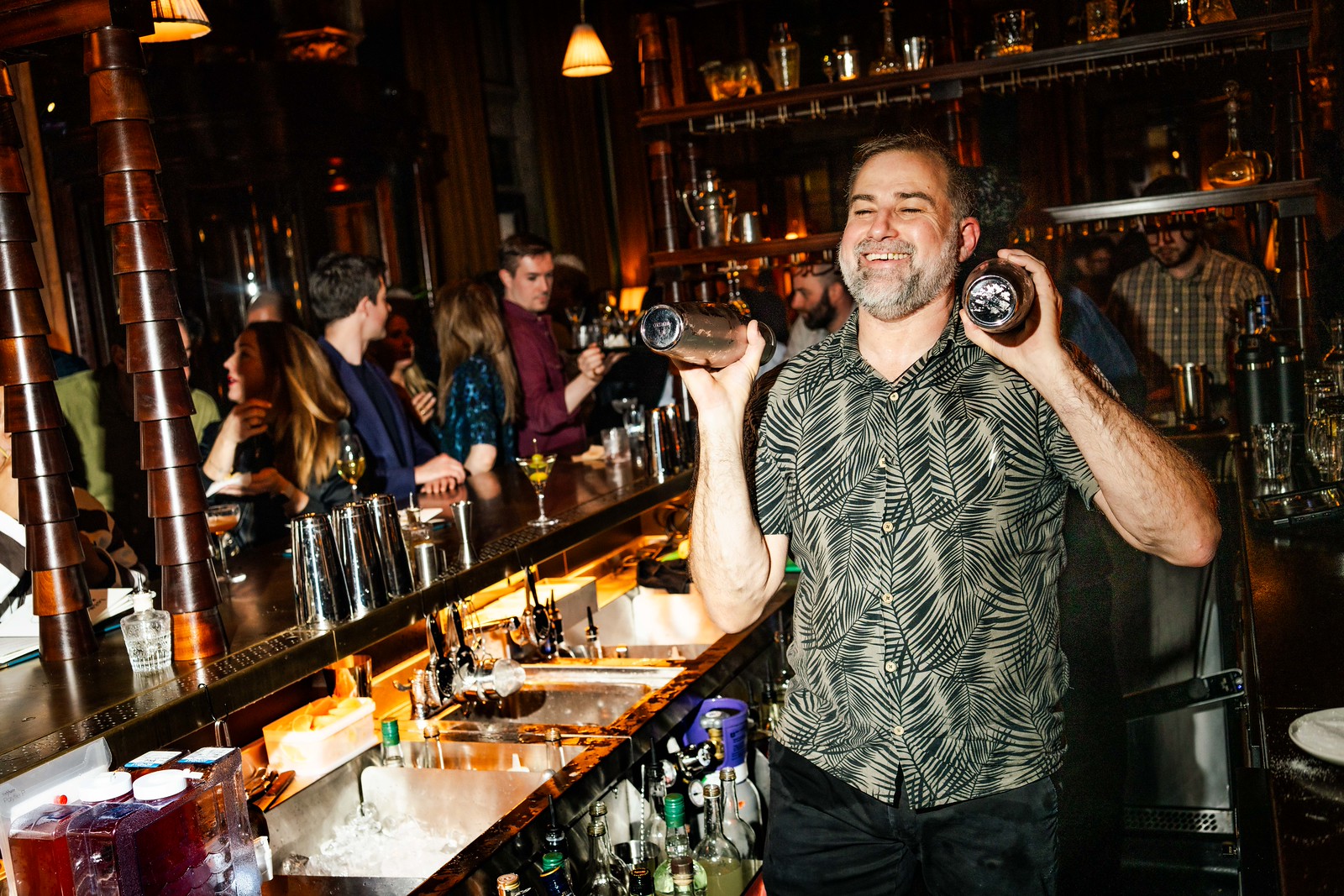 A smiling bartender shaking cocktail shakers in both hands - with a party happening in the bar in the background
