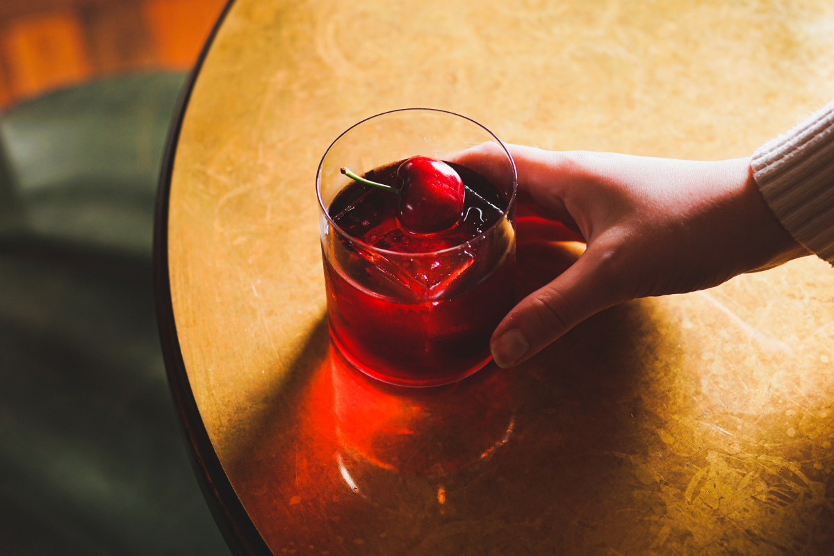 A cherry negroni cocktail being placed on a golden cocktail table by a female hand