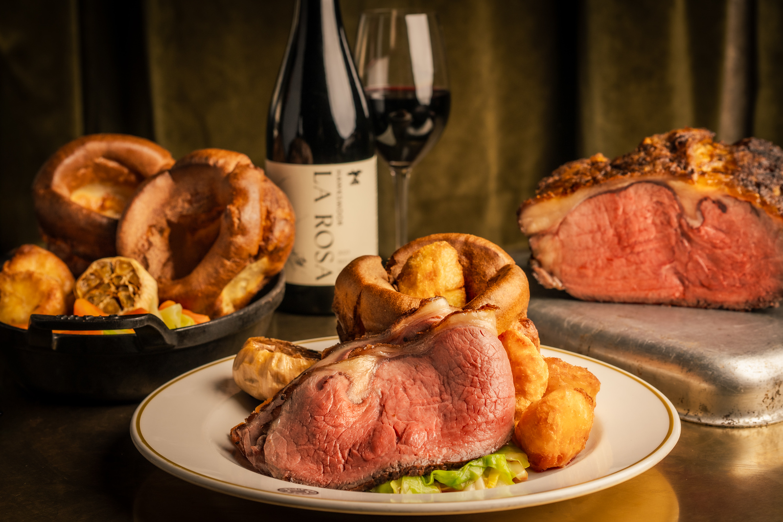 A shot of a sirloin Sunday Roast with all the trimmings in the forefront, on a white place, with a plate piled high with Yorkshire puddings, a bottle of red wine and a joint of beef in the background