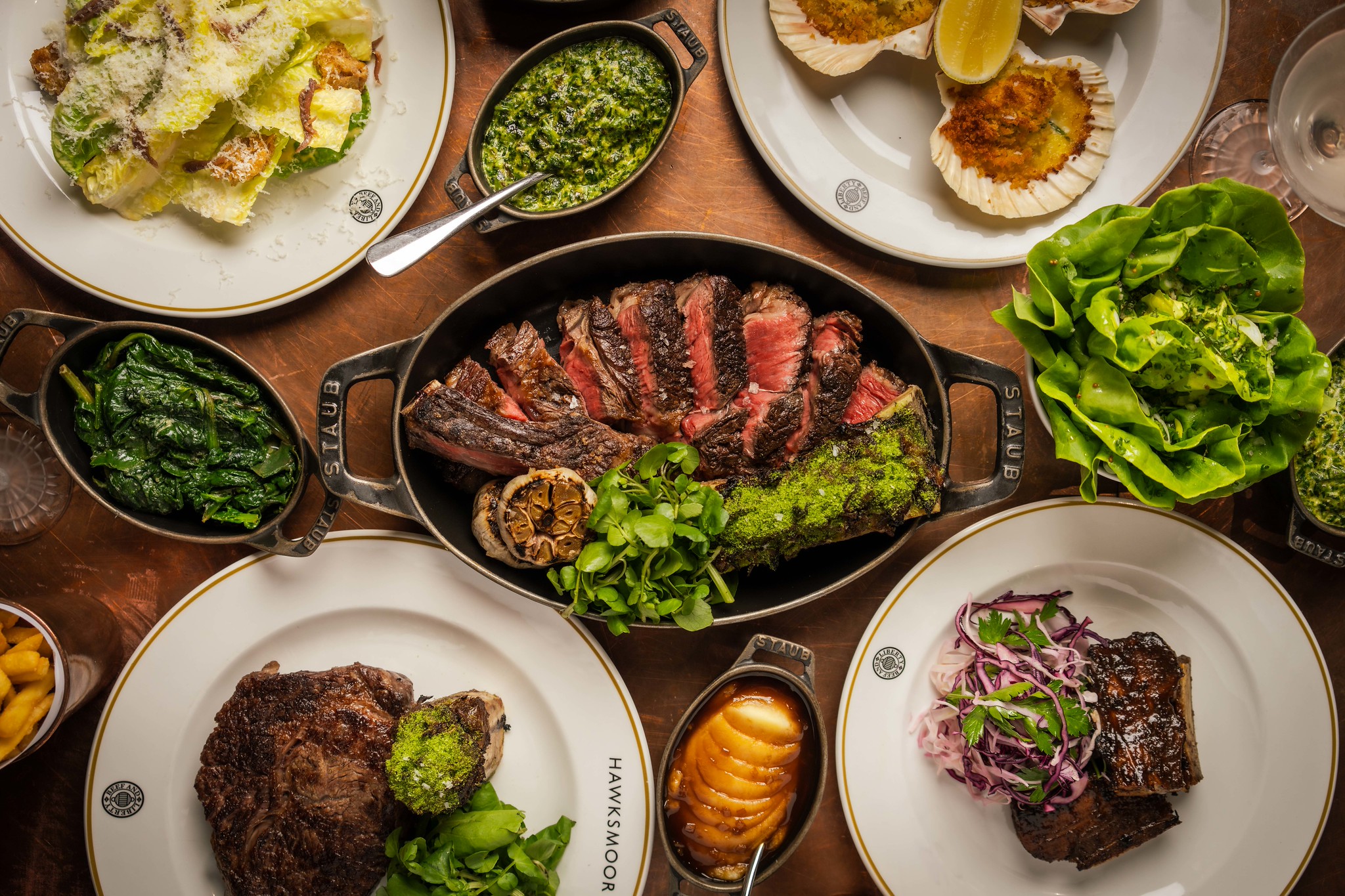 An overhed shot of a table laden with steaks, a sharing steak in the midlle, starters and sides all around