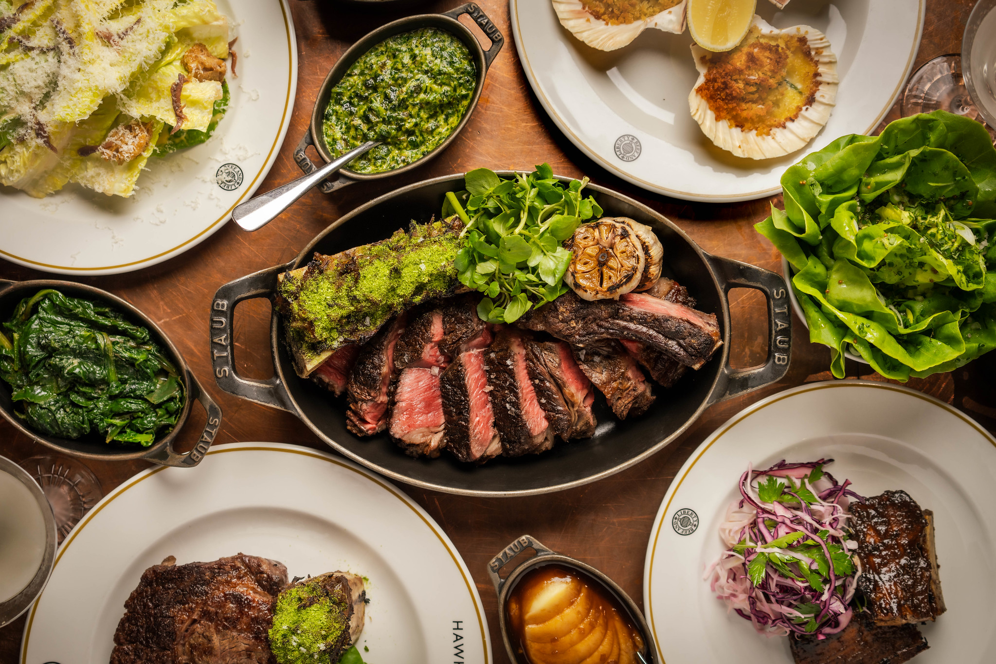 Overhead spread with sliced bone-in steak in a cast-iron dish, ribs, scallops, salads and green sides