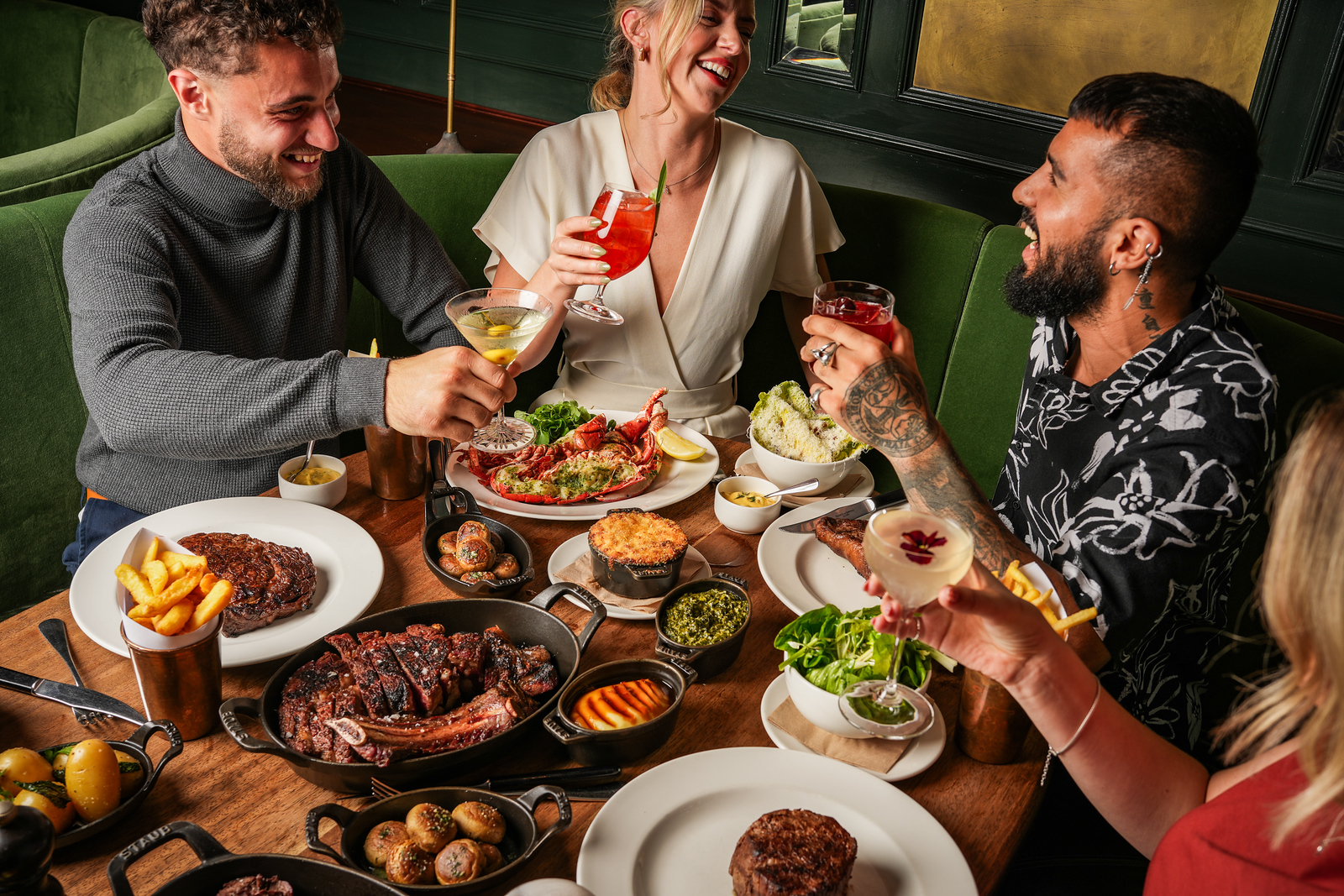 A group of three friends around a table laden with steak, seafood and sides, laughing and raising glasses