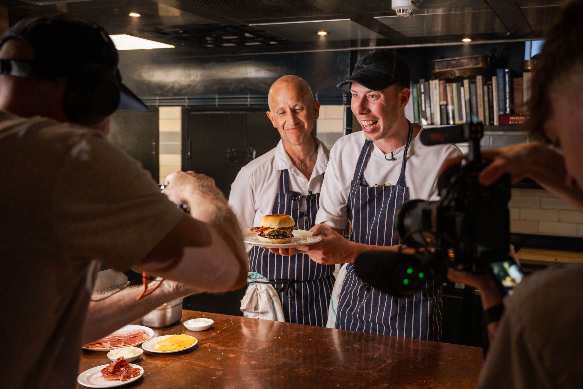Smiling Calfreezy holding up a plate with the Burguur to be shot with a camera, smiling Chef Matt in the background