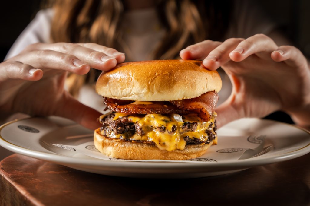 Calfreezy x Hawksmoor 7 Close-up shot of the Burgurr on a white plate, with two hands preparing to pick it up