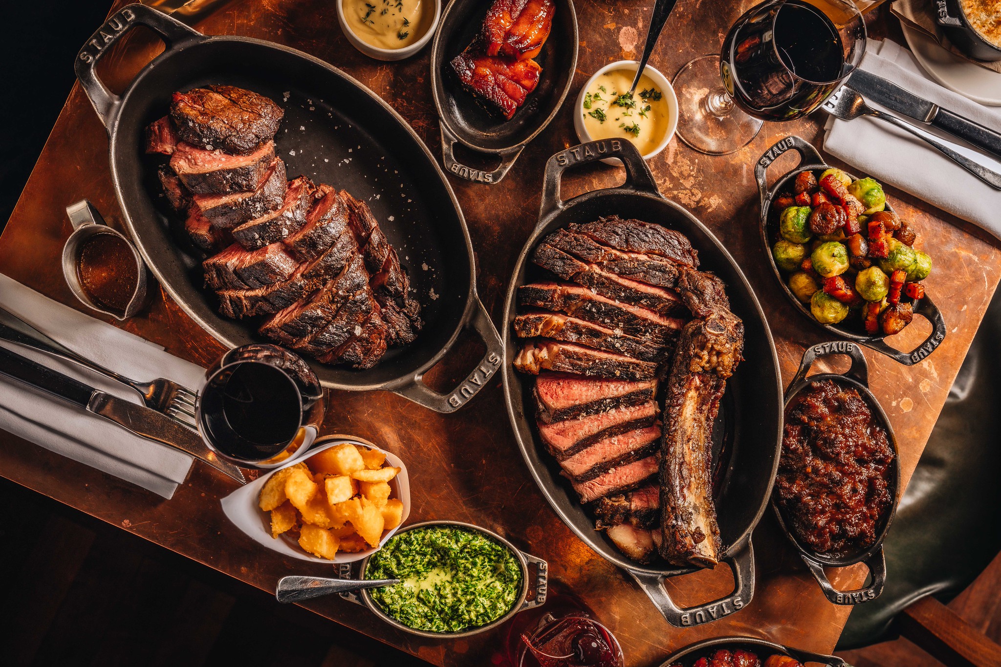 An overhead shot of two cast iron skillets with sharing steaks sliced up, with some sides around on the table