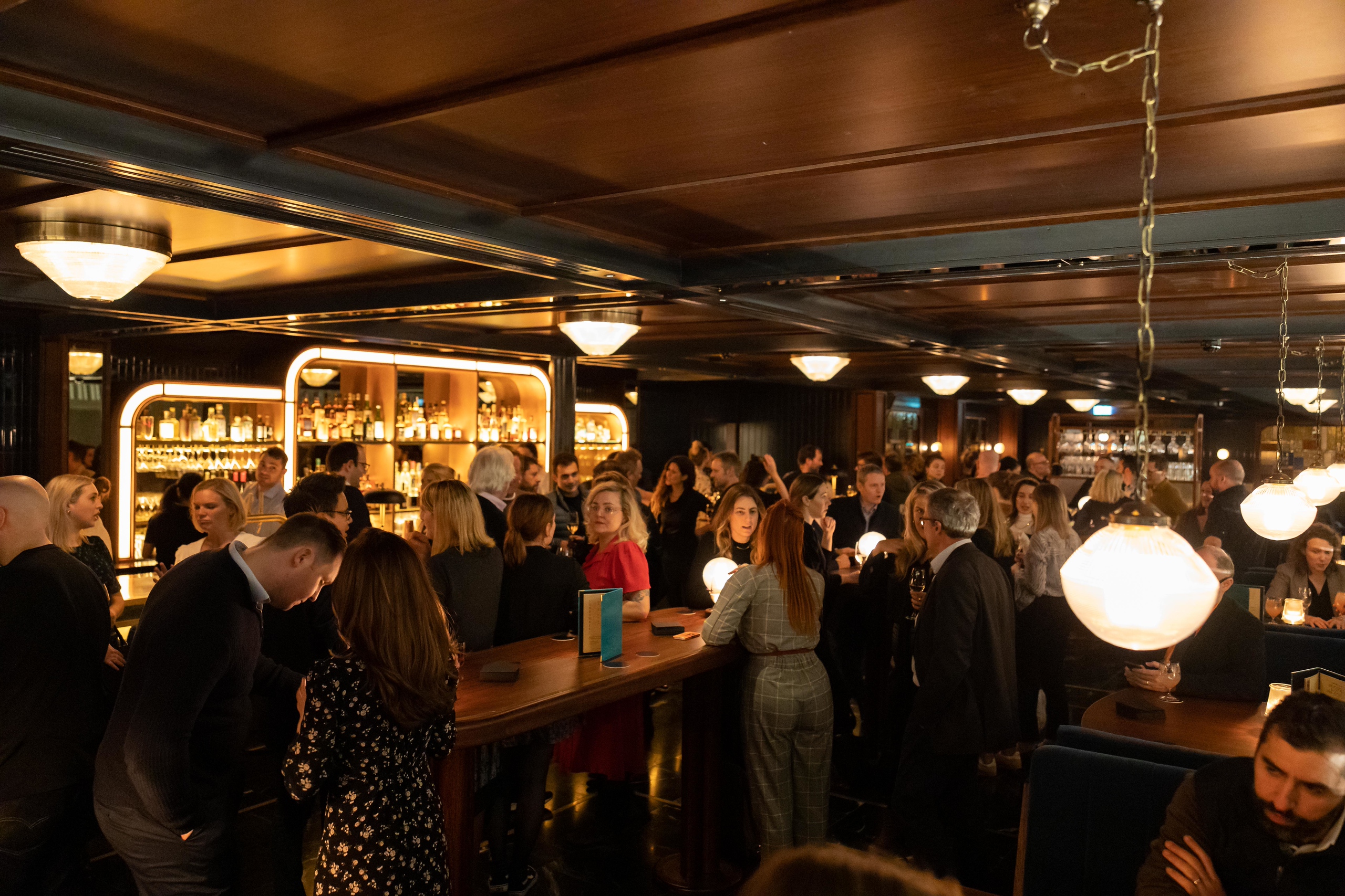 A busy private drinks reception in the bar at Hawksmoor Wood Wharf, with guests standing with drinks in hand around the bar, backlit shelves of bottles behind and warm globe lighting overhead