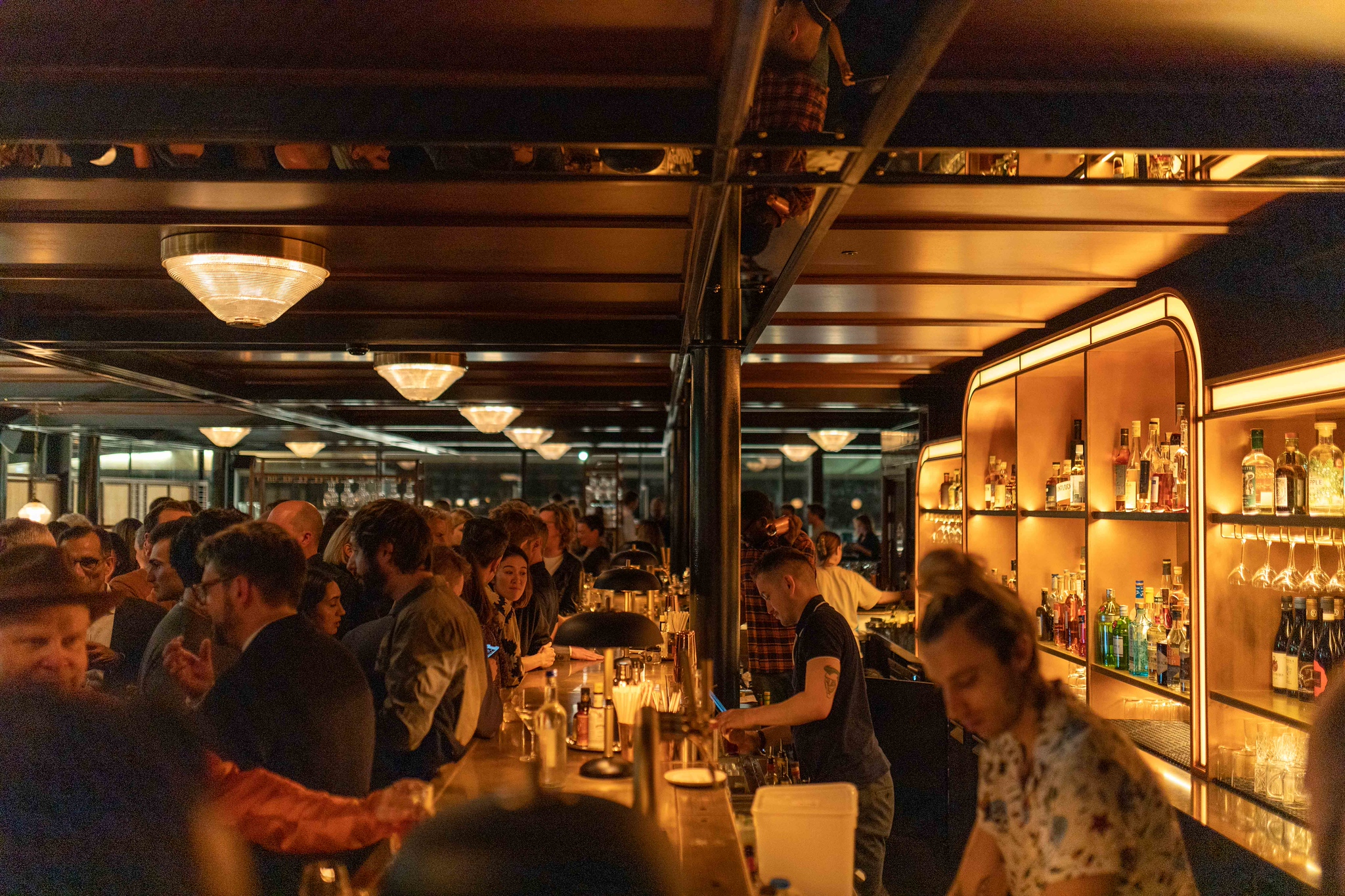 A busy evening at the bar in Hawksmoor Wood Wharf, with guests seated the length of the bar under warm low lighting, bartenders mixing drinks and brass-edged shelves of bottles and glassware lit up behind them
