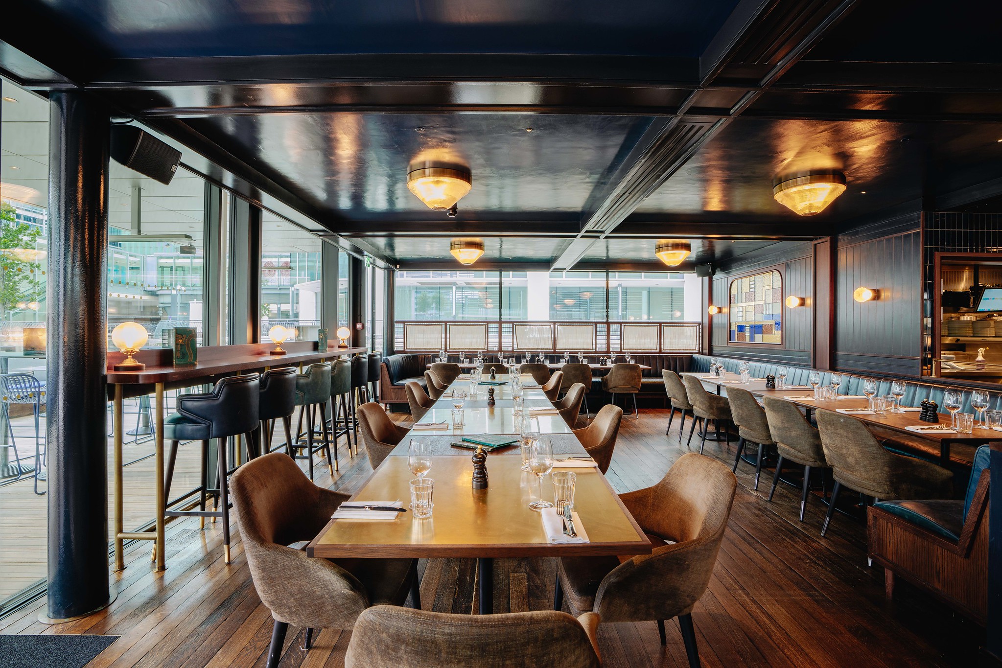 The semi-private dining space in the bar at Hawksmoor Wood Wharf, with two long wooden tables set for a group, leather and velvet armchairs, floor-to-ceiling windows and high counter seating along the window