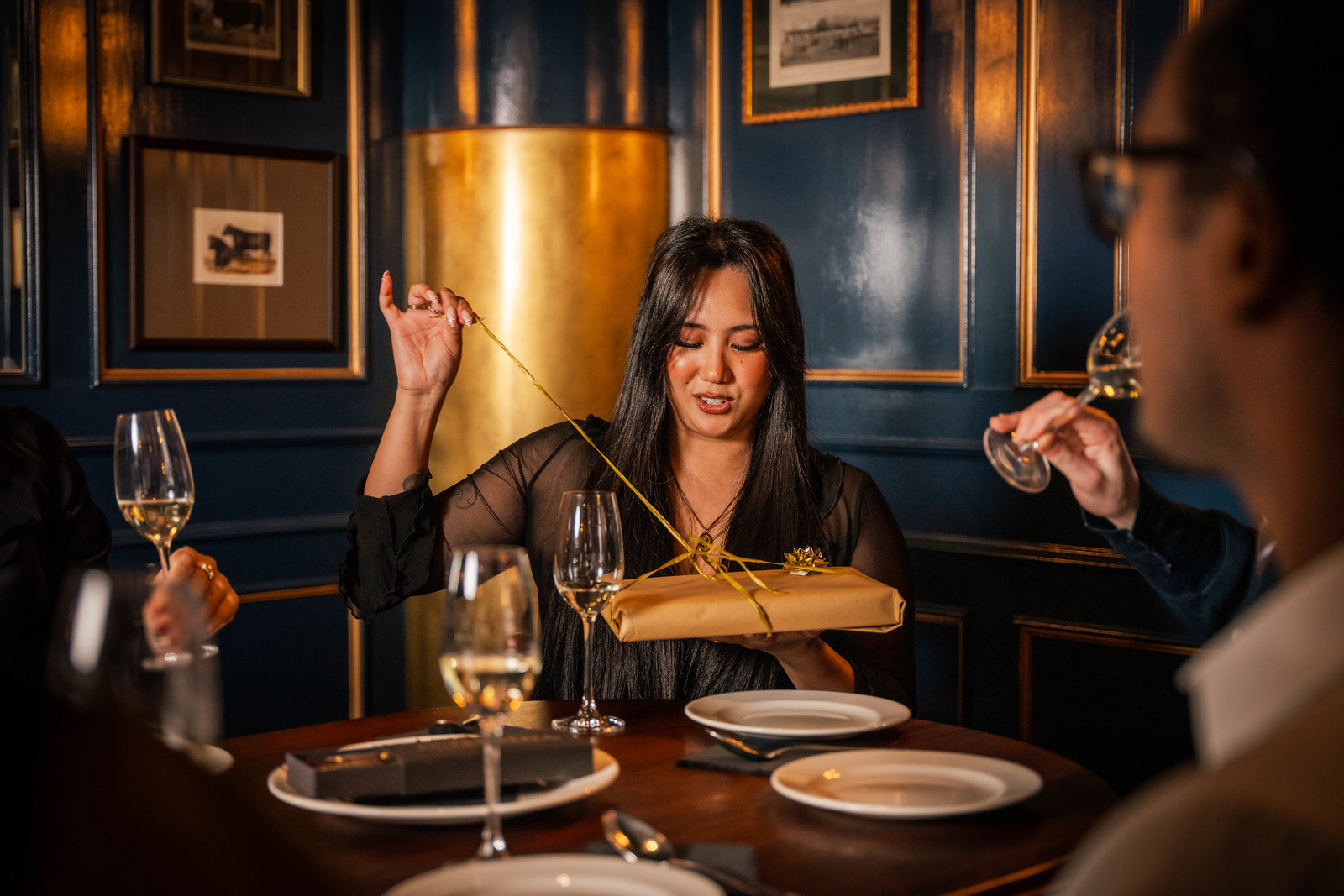 Three guests celebrating a birthday in a wood-panelled Hawksmoor dining room, untying a paper-wrapped gift on a table set with wine glasses