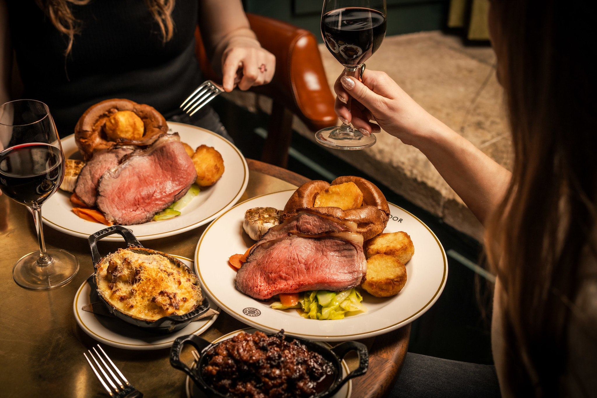 Two plates of pink roast beef with a Yorkshire pudding, roast potatoes and greens, red wine, and side dishes on a dark table.