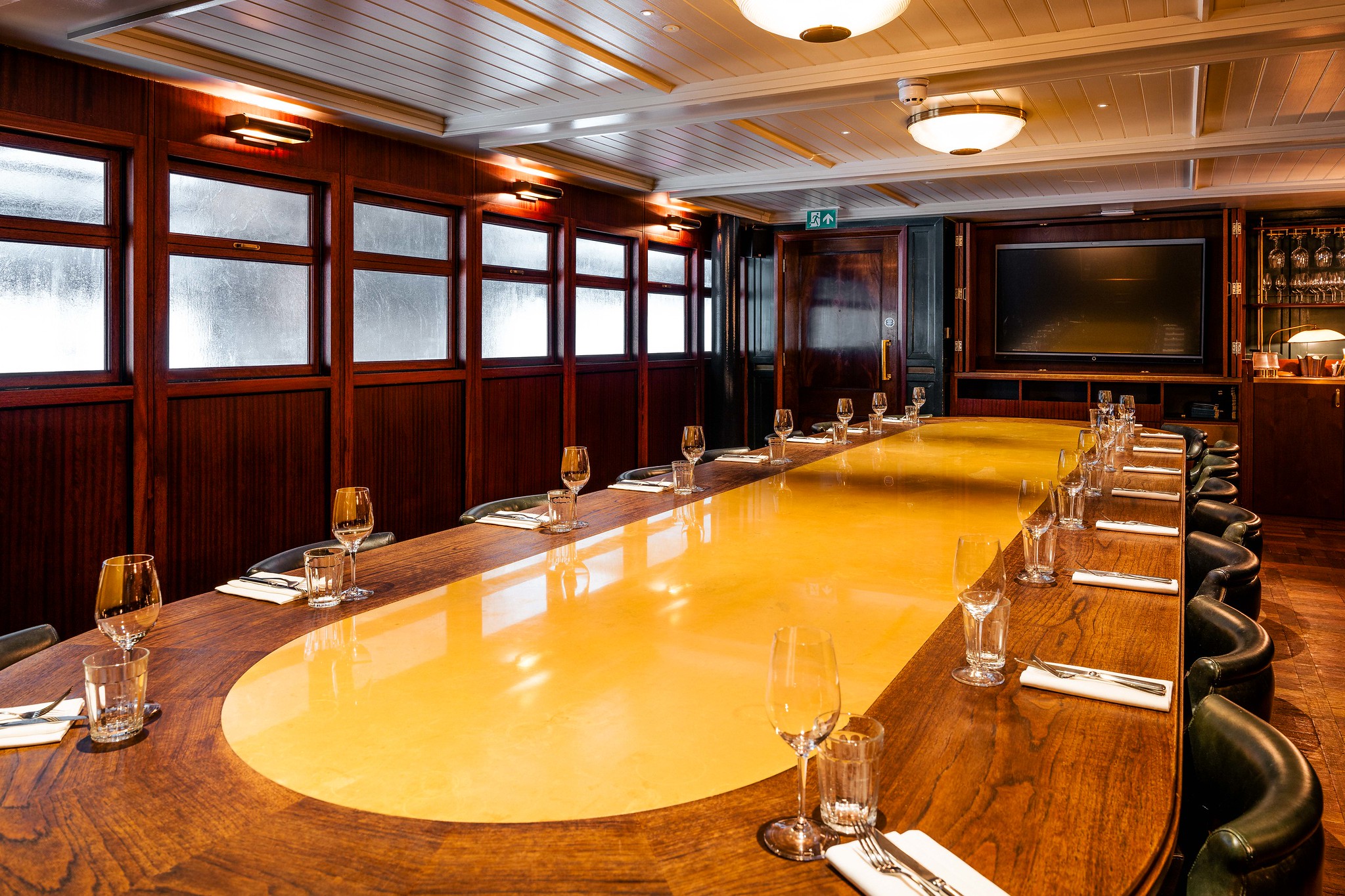 The Queenie Watts private dining room at Hawksmoor Wood Wharf, laid for up to 21 guests around a long oval table, with wood panelling, leather chairs and a wall-mounted screen for presentations