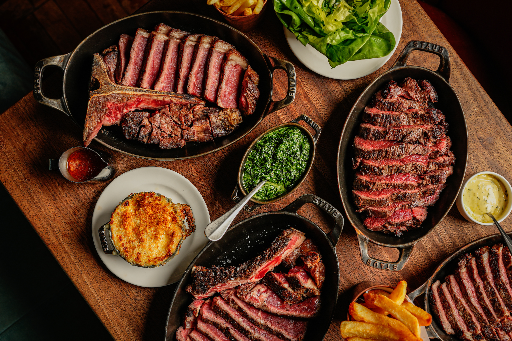 Sharing steaks at Hawksmoor, four cast iron dishes of sliced steak with chips, salad and sauces on a wooden table