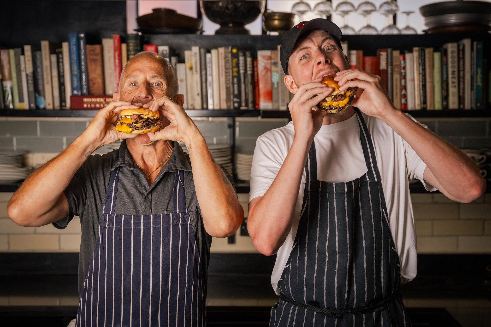 Hawksmoor chef Matt Brown and Calfreezy biting into cheeseburgers in front of a wall of cookbooks