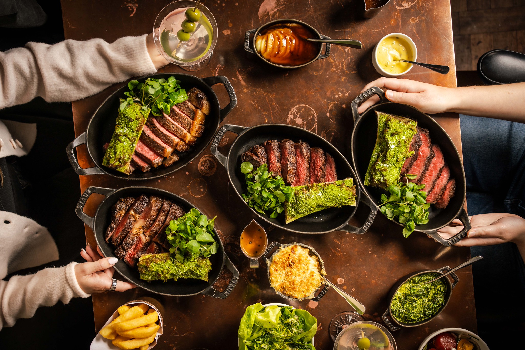 Friends sharing a Hawksmoor steak flight, with sliced steaks in cast iron dishes, chips and sides across the table
