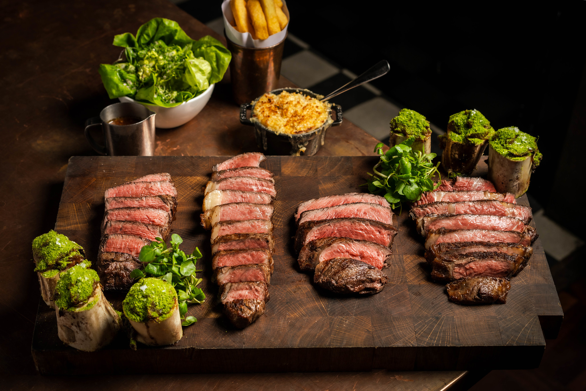 A Hawksmoor steak flight: fillet, sirloin, rump and ribeye sliced and laid out on a wooden board with sides