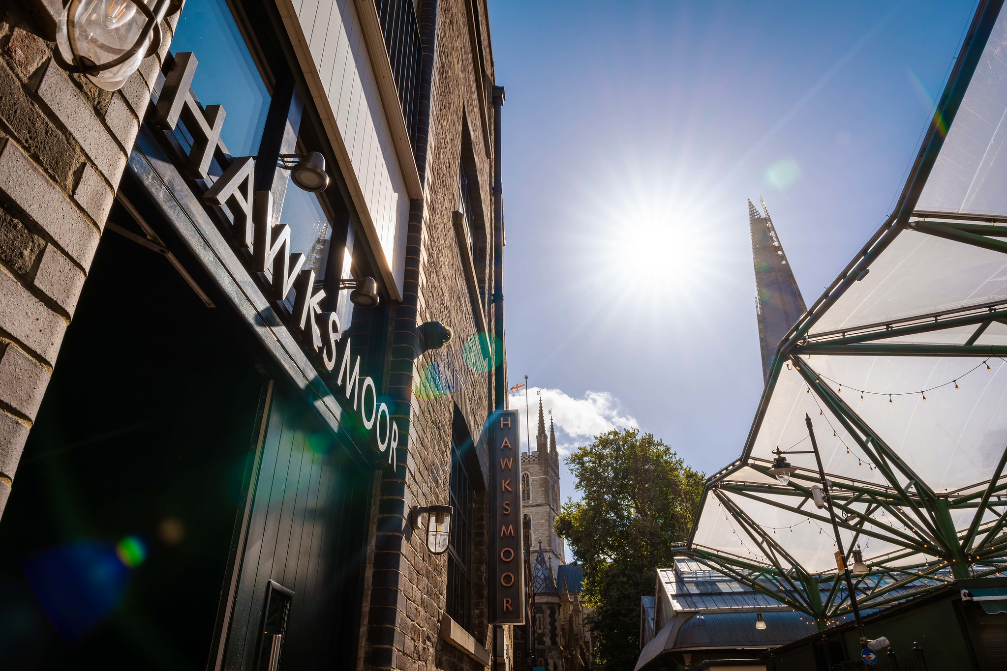 Hawksmoor Borough exterior signage on a dark shopfront, with the Shard, a church tower and the Borough Market canopy behind in bright sunshine