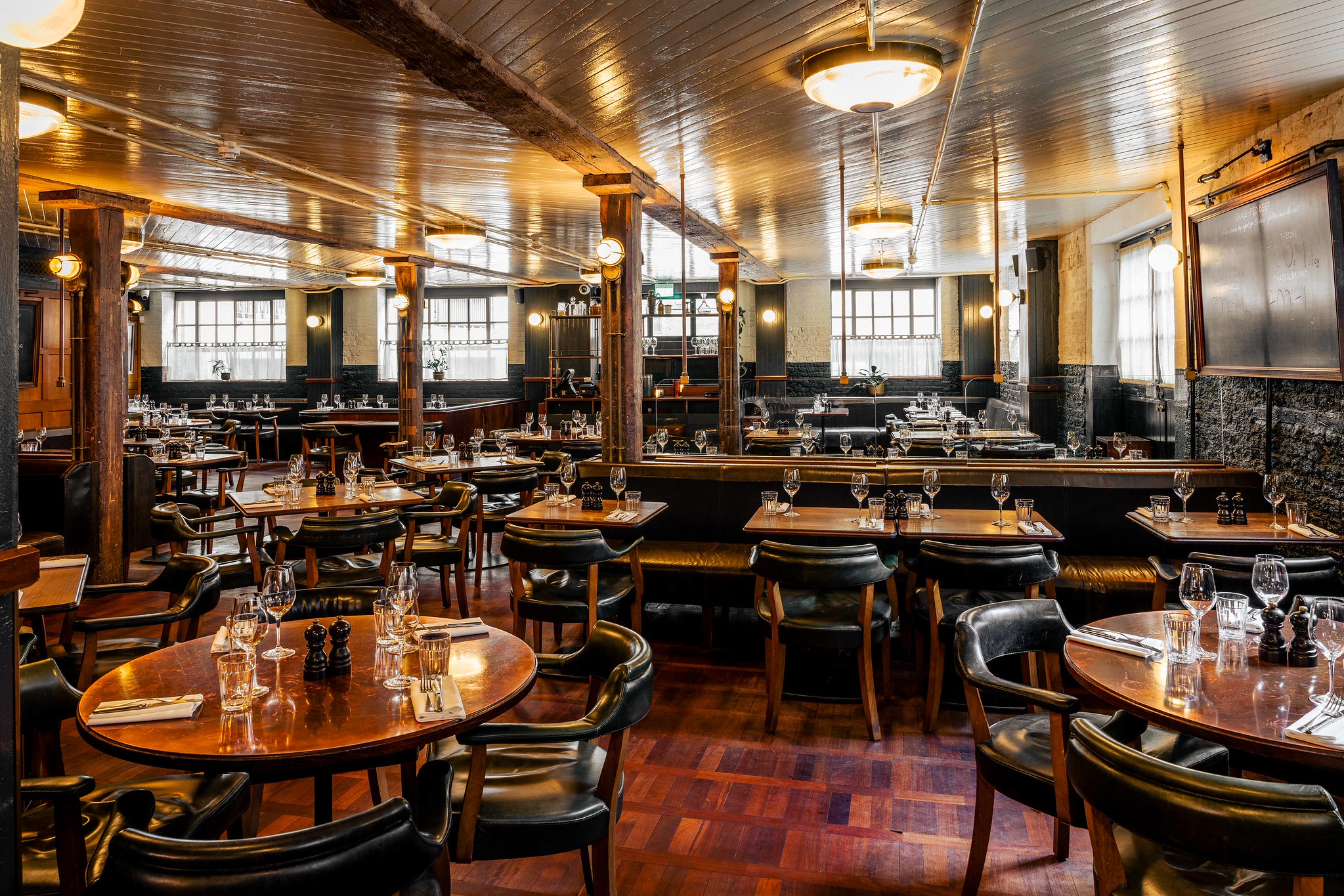 Hawksmoor Borough dining room with timber columns, green leather banquettes, round wooden tables and a terracotta tiled floor, lit by frosted windows